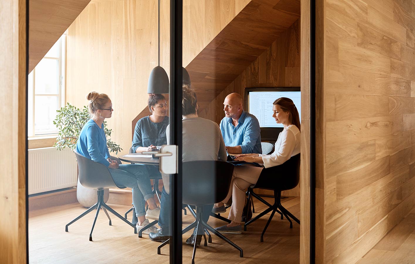 Besprechungraum mit Personen an einen runden Tisch. Die Glastür steht offen.